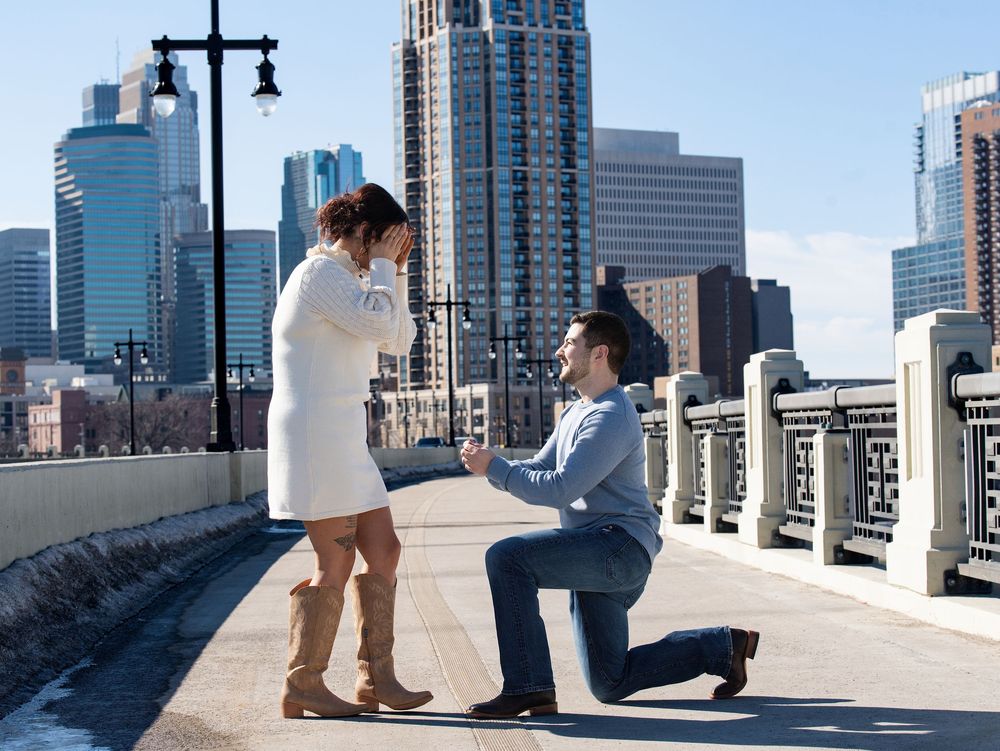 He kneels on a bridge as she covers her face in surprise, skyline behind